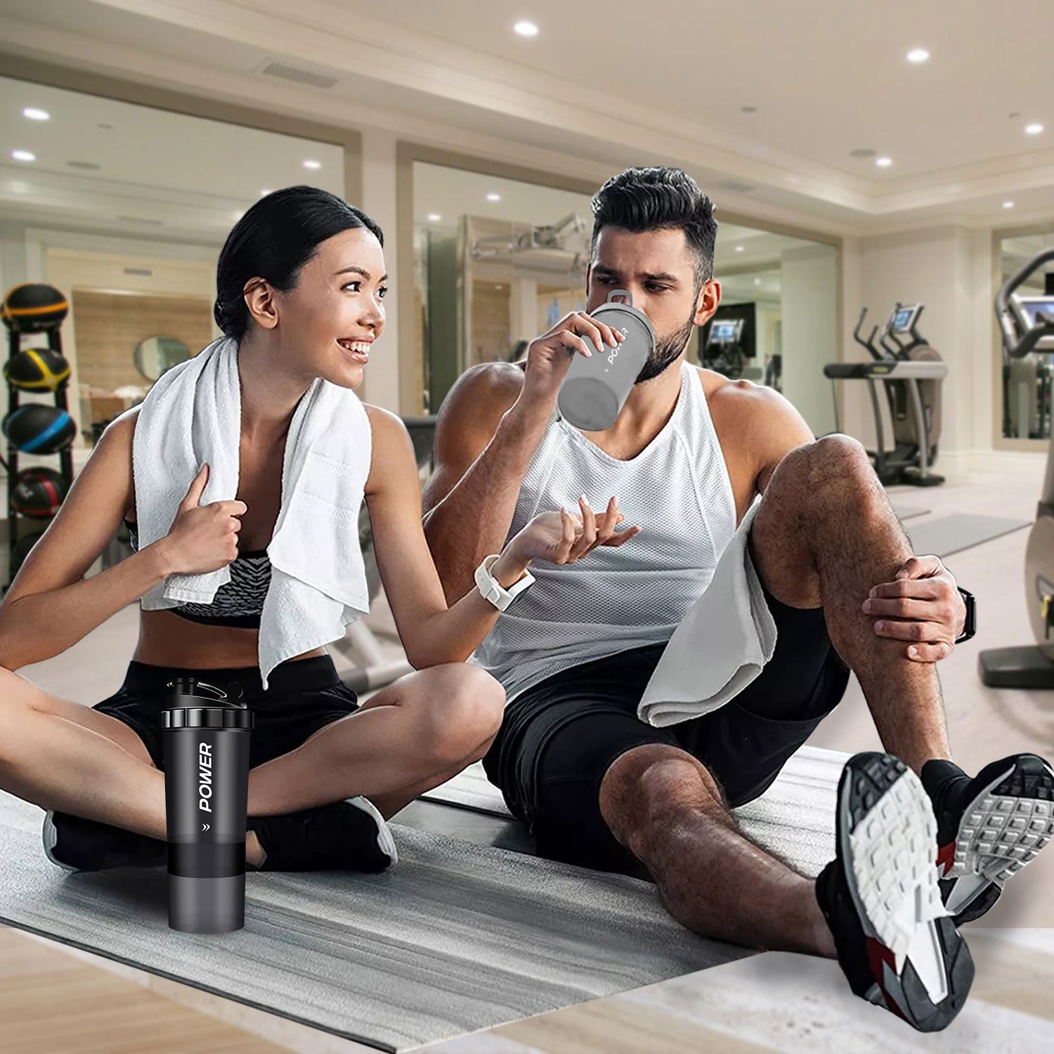 A man and woman sit on the gym floor, surrounded by fitness equipment, discussing their Acuna protein shaker